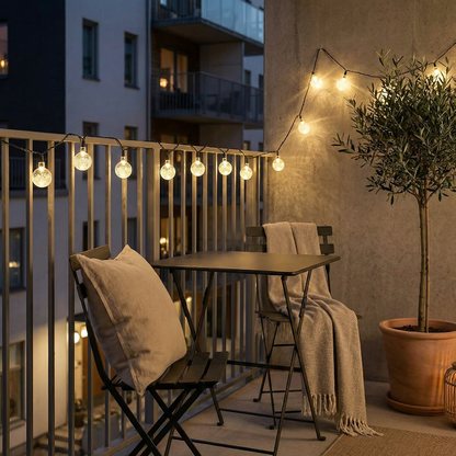 Balcony with string lights, table, chairs, and potted plant at night.