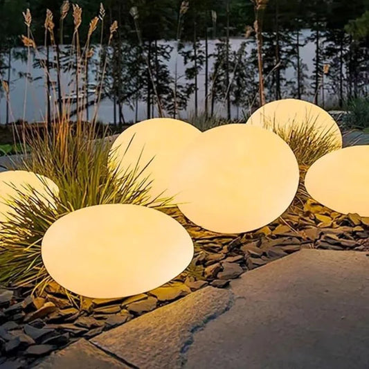 A close-up shot of several large, round, illuminated lights resembling pebble stones, placed among dark rocks and tall grasses in an outdoor garden. The lights are solar-powered and provide a soft, warm glow.