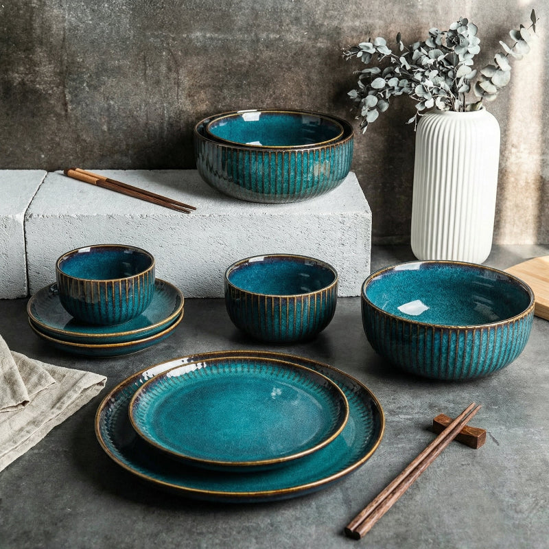 Set of blue ceramic dishes with gold rims on a dark surface, with a vase and plant in the background.