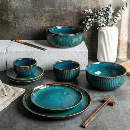 Set of blue ceramic dishes with gold rims on a dark surface, with a vase and plant in the background.