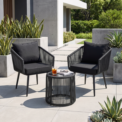 Two black chairs with gray cushions and a small round table on a patio with plants and a building in the background.
