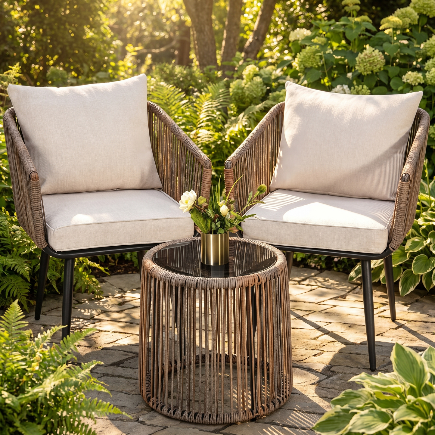 Two wicker chairs with beige cushions and a matching table on a patio surrounded by greenery.