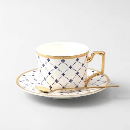 White Amalfi ceramic mug and saucer with blue lattice pattern and gold trim and a gold spoon on a clean off-white background