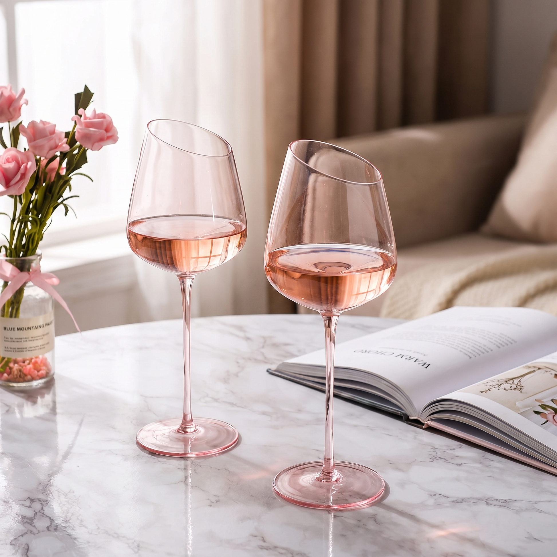 Blush pink wine glasses with rose on a marble table beside a book and flowers