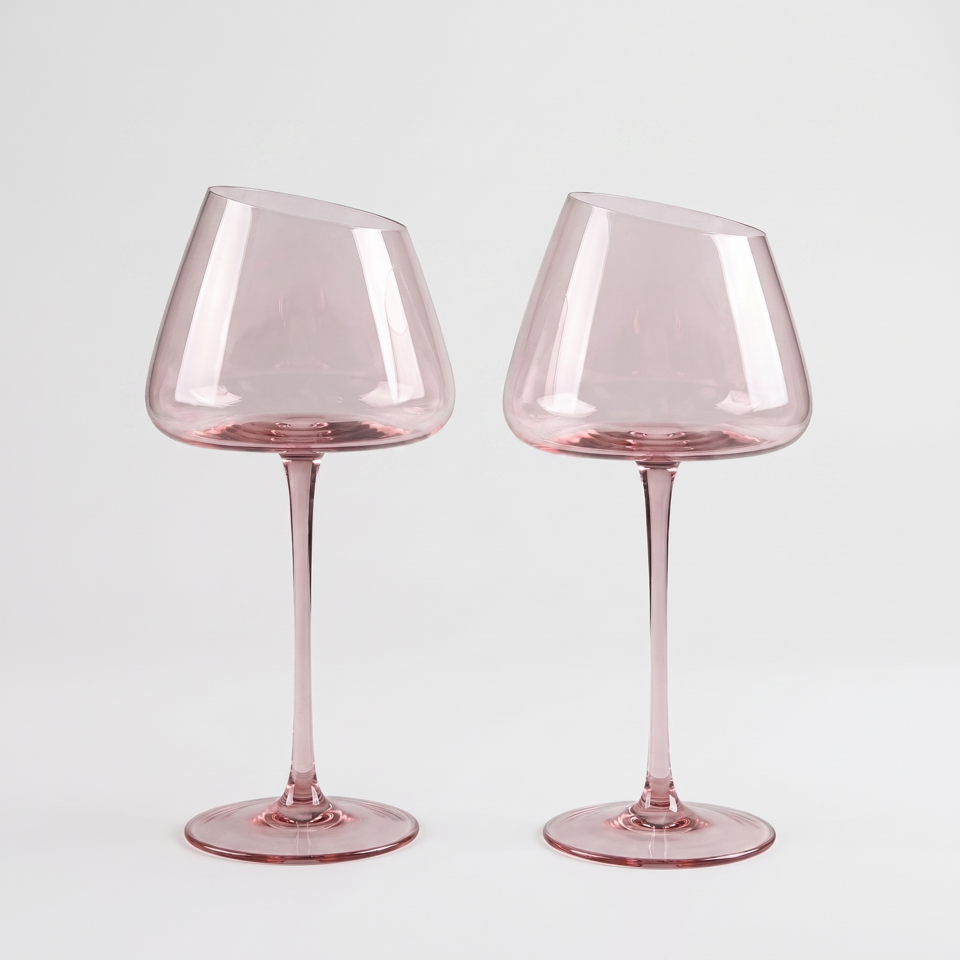 Set of 2 blush pink hand-blown crystal red wine glasses with an angled rim and pink stem on a clean off-white background