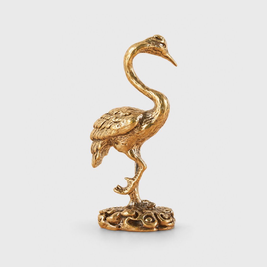 Polished brass crane figurine standing on one leg with folded carved wings on a round openwork base, on a plain light gray background