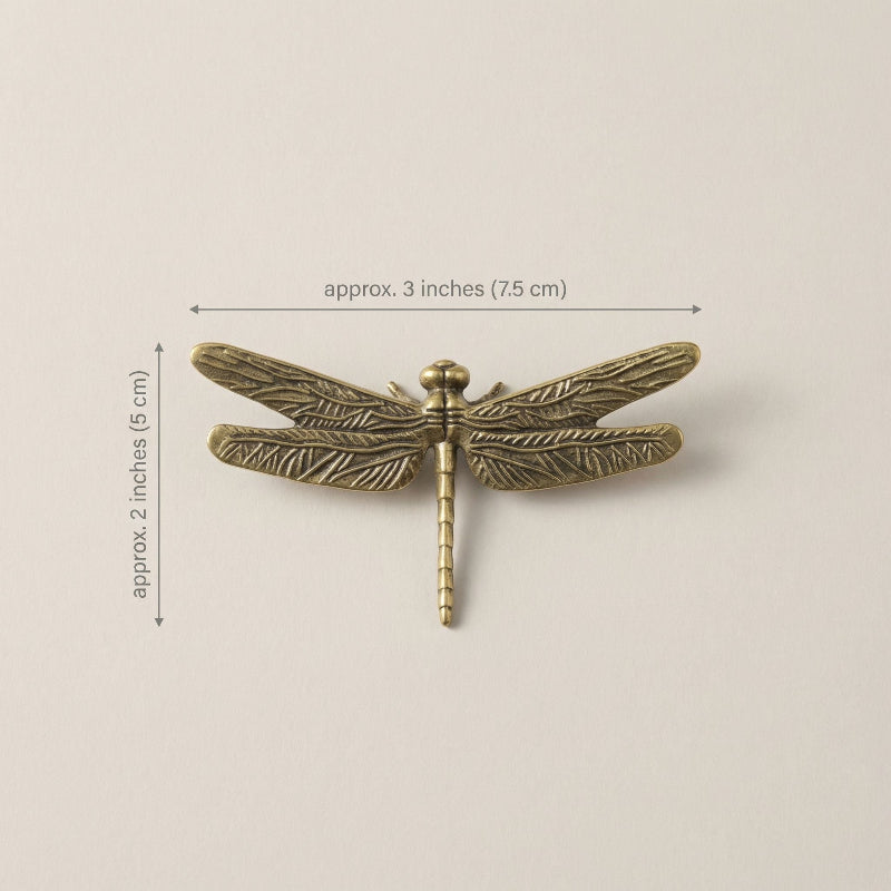Bronze dragonfly-shaped object with measurements on a beige background