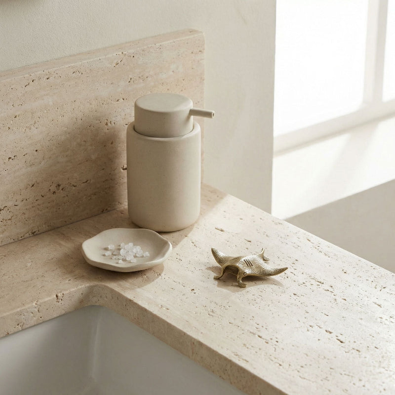 Beige soap dispenser on a bathroom counter with a brass mantaray and small dish.