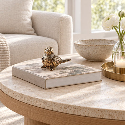 Antiqued brass sparrow figurine perched on a linen-covered book on a round stone coffee table beside a speckled bowl, white flowers and a lit candle