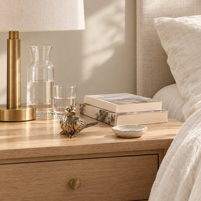Brass sparrow figurine on a light oak nightstand beside a brass lamp, a glass carafe of water and two stacked books