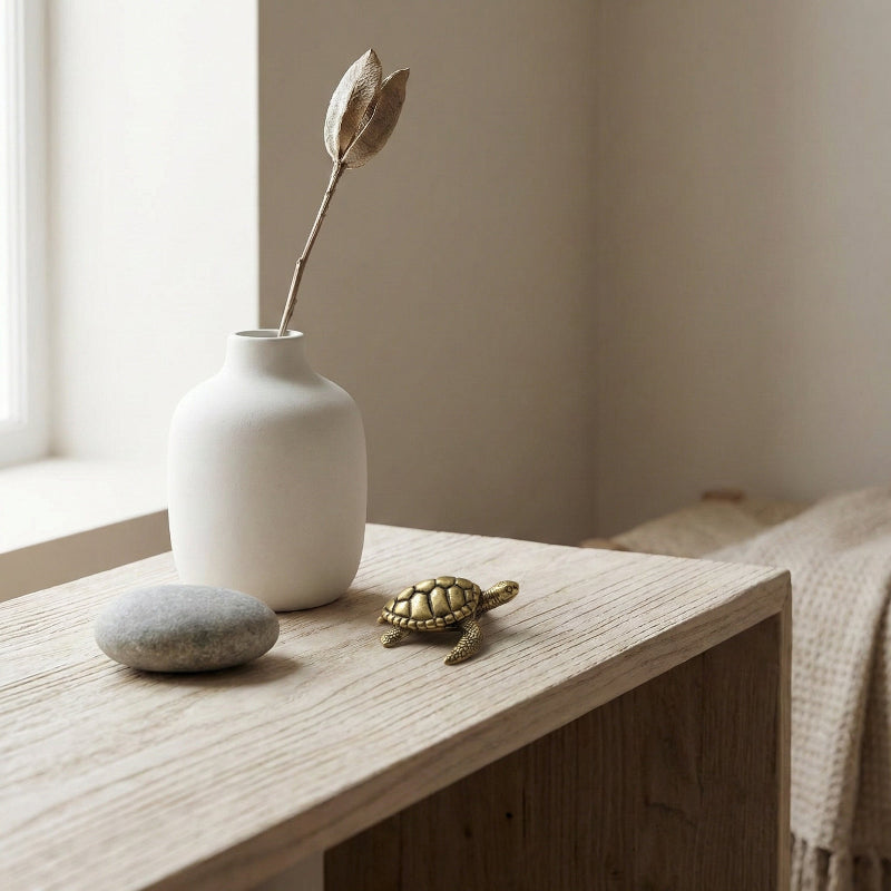 White vase with a dried flower, stone, and small turtle figurine on a wooden surface near a window.