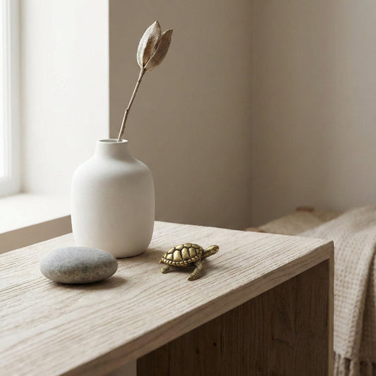 White vase with a dried flower, stone, and small turtle figurine on a wooden surface near a window.