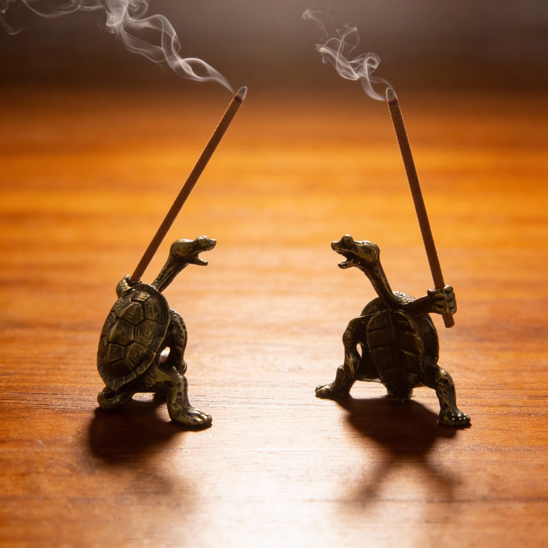 Two turtle-shaped incense holders with smoking sticks on a wooden surface.