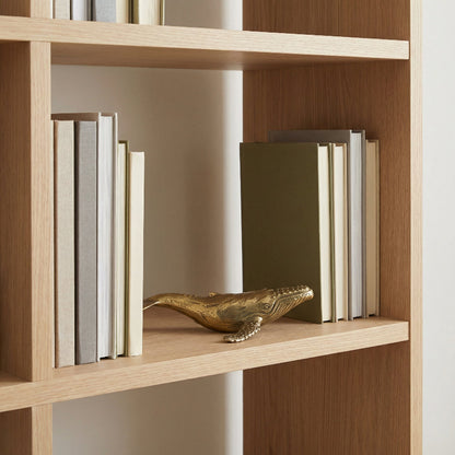 Wooden bookshelf with books and a decorative brass whale on a neutral background.
