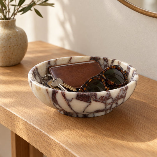 Shallow white marble bowl with burgundy veining on a light wood console holding keys, a brown wallet and tortoiseshell sunglasses, with a vase of leafy stems and a round mirror edge behind
