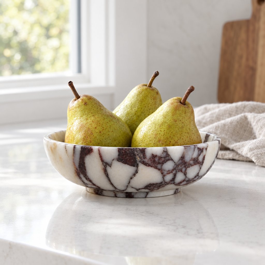 Shallow white marble bowl with burgundy veining on a white kitchen counter holding three green pears, with a window, a wooden board and a linen cloth behind