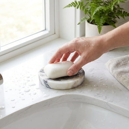 Hand placing soap on a marble soap dish by a window with a plant in the background.