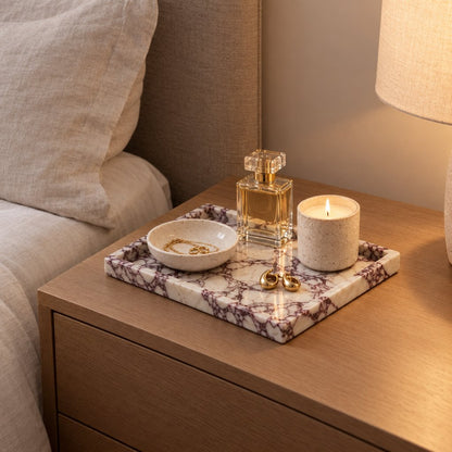 White marble tray with burgundy veining on a bedside table holding a perfume bottle, a lit candle, a jewelry dish and gold earrings