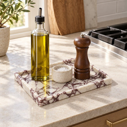 White marble tray with burgundy veining on a kitchen counter holding a glass cruet of olive oil, a stoneware salt cellar and a wooden pepper mill