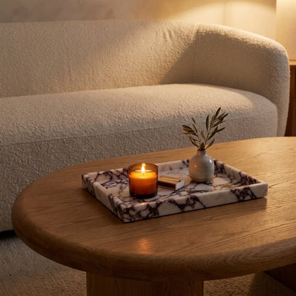 White marble tray with burgundy veining on a coffee table holding a lit candle in an amber jar, a brass matchbox and a small vase with an olive branch