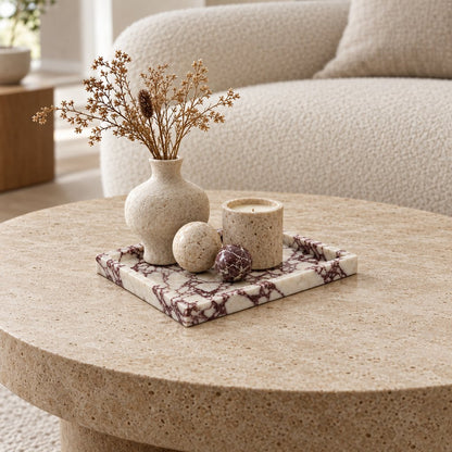 White marble tray with burgundy veining on a travertine coffee table holding a beige vase of dried stems, two stone spheres and a candle holder
