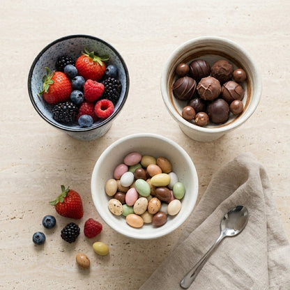 Overhead view of three ceramic dessert bowls holding mixed berries, chocolate truffles and candy-coated almonds on a travertine surface