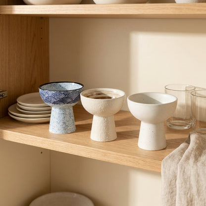 Three empty ceramic pedestal dessert bowls in blue wave pattern, cream with a brown swirl inside and plain white on an oak shelf