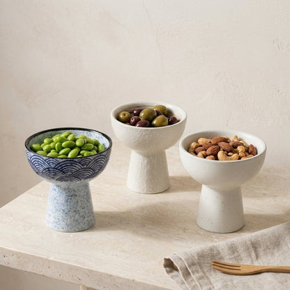Three ceramic pedestal dessert bowls holding edamame, olives and mixed nuts on a travertine table