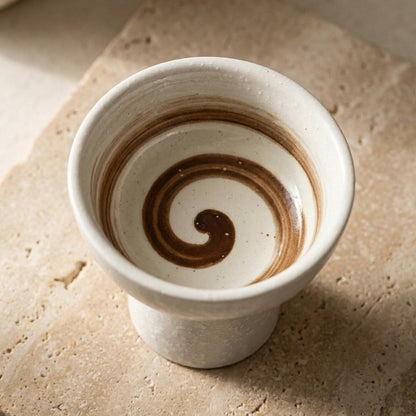 Close-up of an empty cream ceramic pedestal dessert bowl with a brown spiral swirl glaze inside on a travertine slab