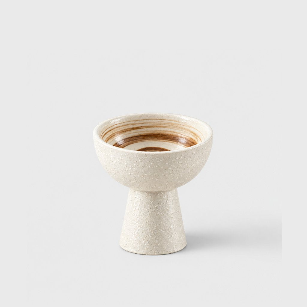 Single cream pitted ceramic pedestal dessert bowl with a brown swirl glaze inside on a light grey background