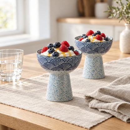 Two blue and white wave pattern ceramic pedestal dessert bowls of yogurt with berries and honey on an oak table with a linen runner