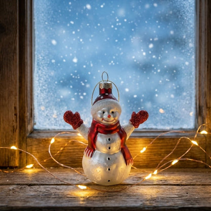 Glass snowman ornament on windowsill with string lights, snow outside