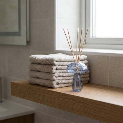 Blue glass mushroom vase used as a reed diffuser holder on a bathroom shelf next to folded towels and a window