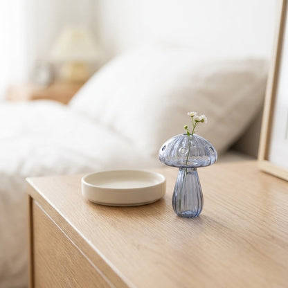 Blue glass mushroom vase holding small white daisies on a bedside table with a mirror and bed pillows blurred behind it