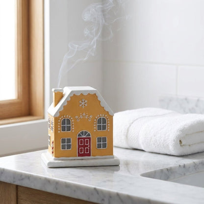 Gingerbread-style cottage incense burner with smoke rising from the chimney, on a marble bathroom counter beside a folded white towel