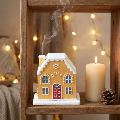Gingerbread-style cottage incense burner with smoke rising from the chimney, on a styled shelf beside a lit pillar candle and warm string lights