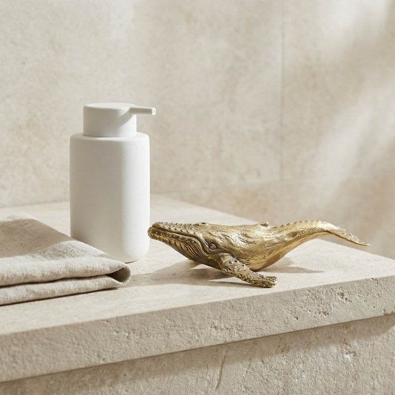 White soap dispenser and gold whale figurine on a stone surface with a neutral background