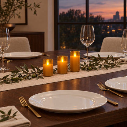 Three amber tinted flameless candles on a wood dining table with an olive branch runner, white plates, gold cutlery and a wine glass, with a dusk sky behind
