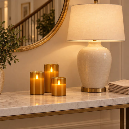 Three amber tinted flameless candles on a marble console beside a white ceramic table lamp, folded napkins and a gold-framed round mirror