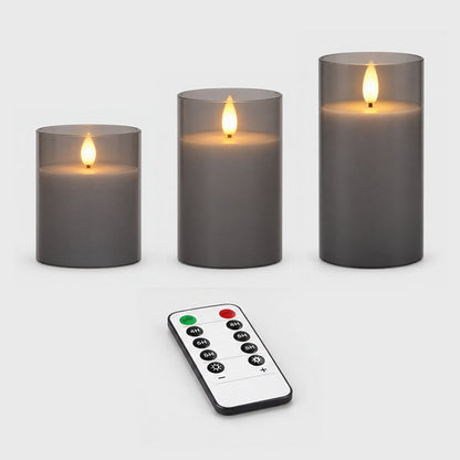 Three grey tinted flameless candles of different heights with a remote control on a plain light background