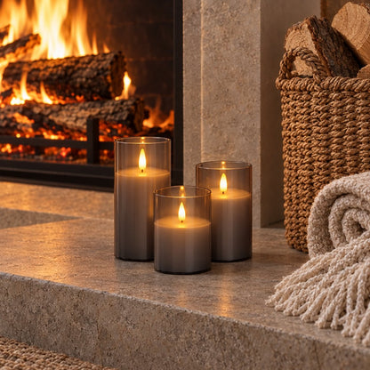 Three grey tinted flameless candles on a stone hearth beside a lit fireplace, a basket of logs and a folded fringed throw