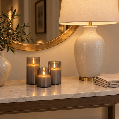 Three grey tinted flameless candles on a marble console beside a white ceramic table lamp, folded napkins and a gold-framed round mirror