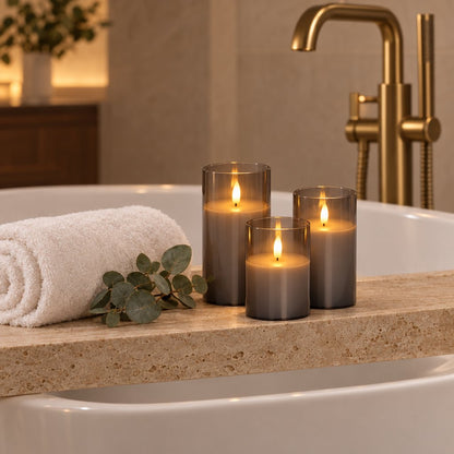 Four grey tinted flameless candles on the stone ledge of a bathtub beside a rolled white towel and eucalyptus leaves, with a gold floor-mounted faucet