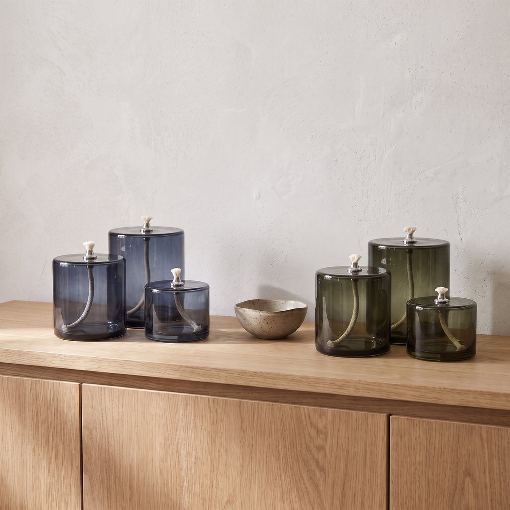 Three smoky blue-grey and three smoky olive green glass oil candles with white wicks on a light wood sideboard beside a small metal bowl