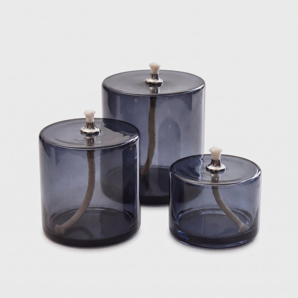 Three smoky blue-grey glass oil candles in small, medium and large sizes with white wicks and silver-toned metal caps on a light grey background