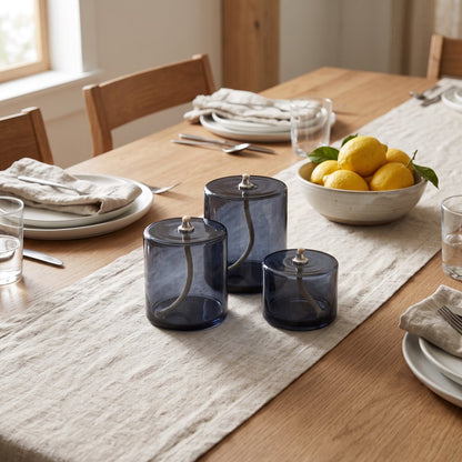 Three smoky blue-grey glass oil candles on a cream cloth table runner on a wooden dining table with a bowl of lemons and place settings