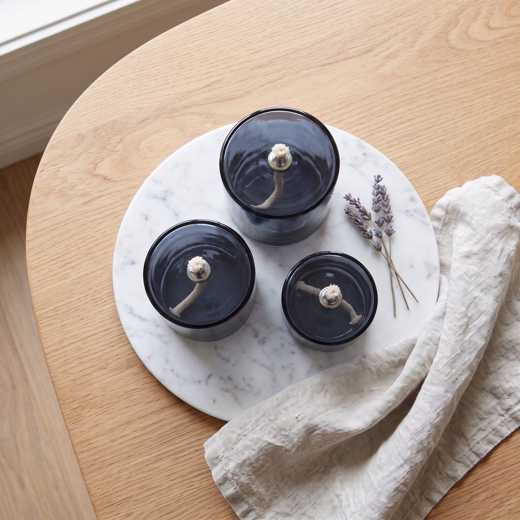 Overhead view of three smoky blue-grey glass oil candles on a round white marbled tray with a cream cloth napkin and lavender sprigs on a wooden table