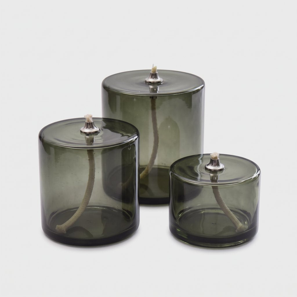 Three smoky olive green glass oil candles in small, medium and large sizes with white wicks and silver-toned metal caps on a light grey background