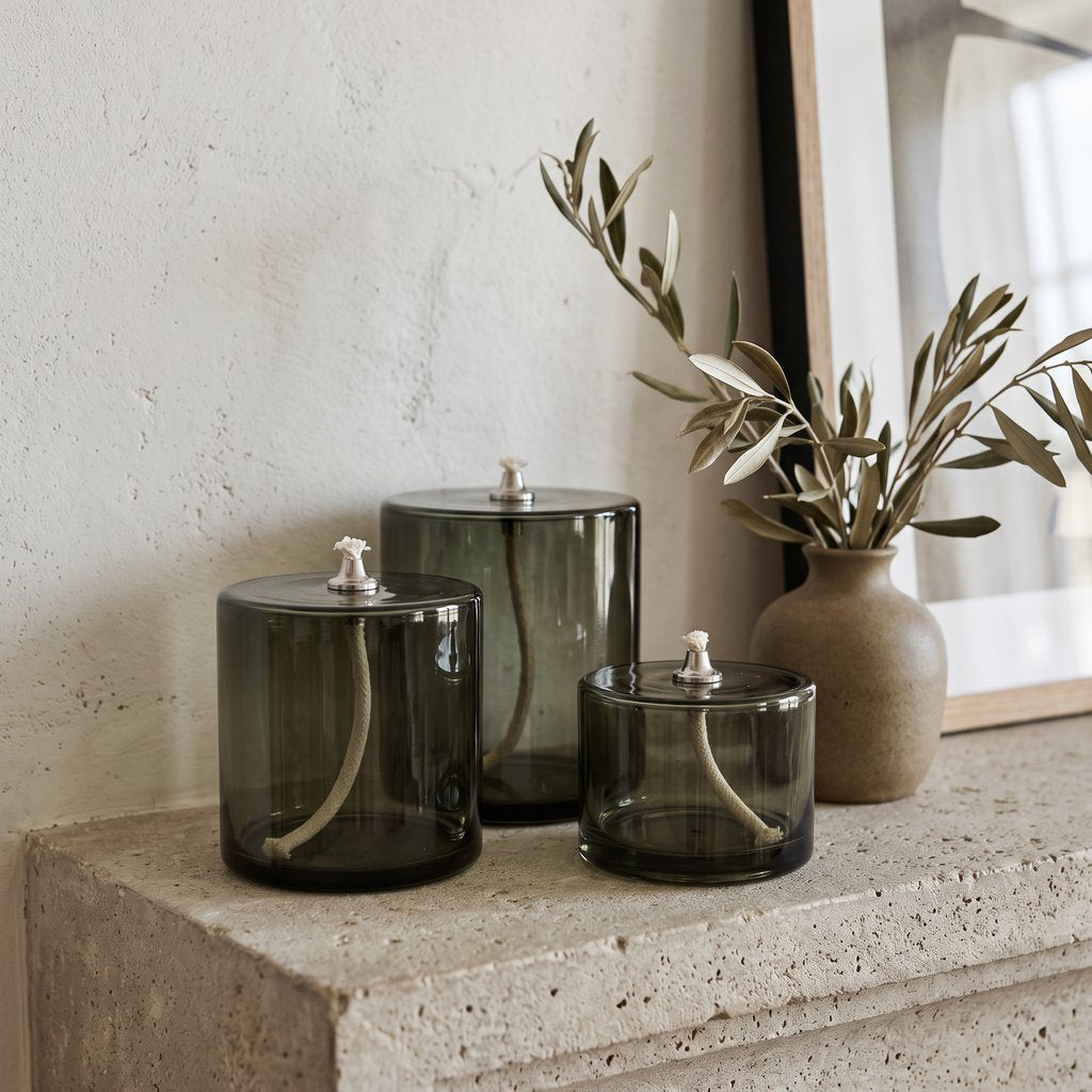 Three smoky olive green glass oil candles on a rough stone ledge beside a leafy branch in a brown vase and a leaning framed picture