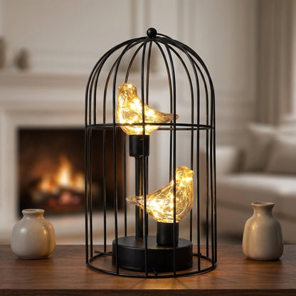 Two Birds illuminated birdcage on a table in front of a lit fireplace flanked by ceramic vases